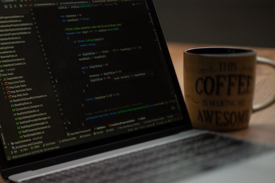 A close-up image of a laptop on a wooden table displaying lines of code on the screen, with a ceramic mug positioned behind the laptop to the right. The mug has a beige background with black text that