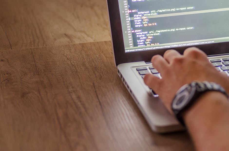 A close-up image of a laptop on a wooden table displaying lines of code on the screen, with a ceramic mug positioned behind the laptop to the right. The mug has a beige background with black text that