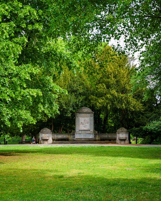A well-maintained public garden near Pinner Memorial Park features a large, lush green lawn with a variety of mature trees, including broad-leaved deciduous trees with vibrant green foliage and conife